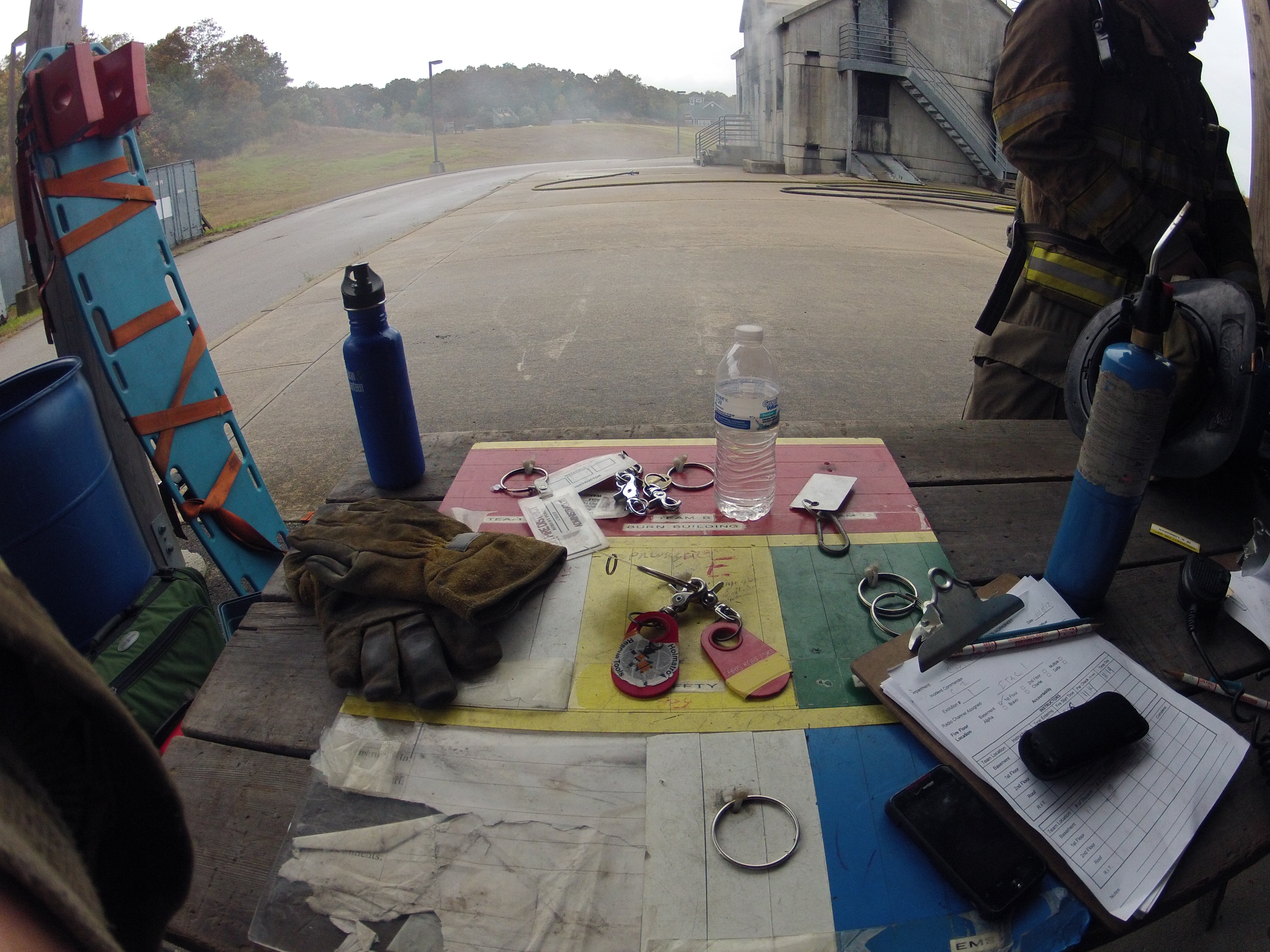 a view of items located on a table where incident managers regularly established a command post during evolutions at the training center. Items include an accountability board, firefighter accountability tags, a clipboard containing check lists, guidelines, and tracking charts, a pair of firefighting gloves, two water bottles, a torch, a cell phone, pencils, and a radio.