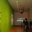 Photograph of a lime green wall on the left with the exhibition title and people walking around the gallery.