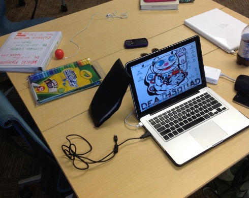 Photograph of a classroom desk with laptop, binder, markers, and other multimodal composing equipment