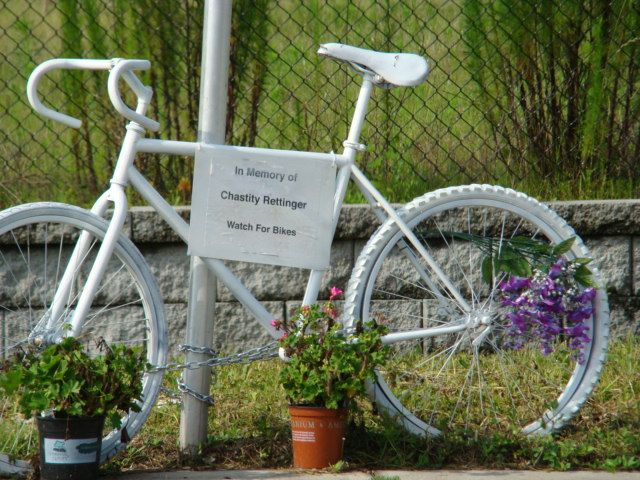 white-painted ghost bike with sign reading 'in memory of Chastity Rettinger; watch for bikes' and live flowers decorating