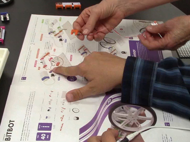 print instructions are open on a table; a male hand points at a figure in the instructions; female hands hold littleBits over the instructions
