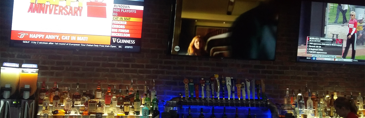 A bank of three large screen televisions mounted about a public bar in Lubbock, Texas. Two are tuned to sports programming and one to a drama. None of the screens are showing captions.