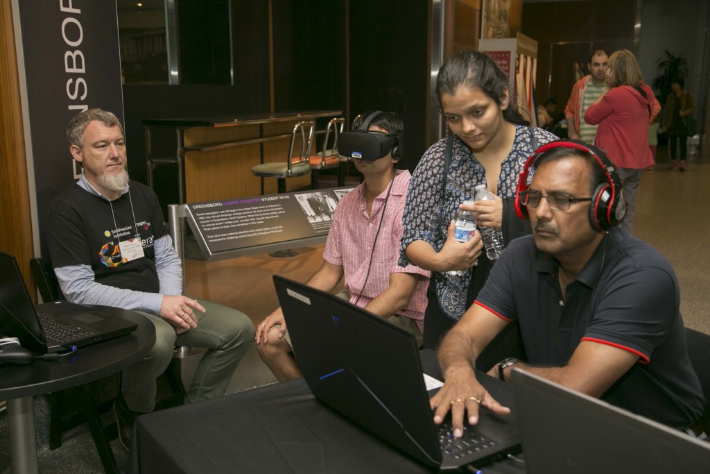 four people around a laptop; one wears headphones and another is wearing a VR headset