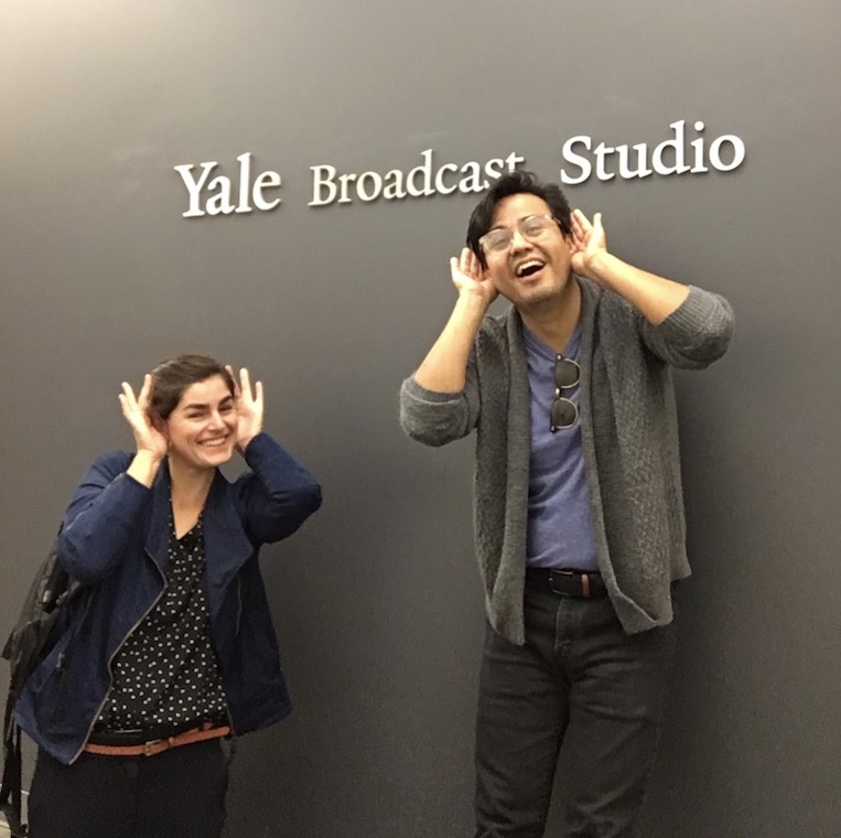 Sylvia Ryerson and Luis Luna stand in front of the the Yale Broadcasting Studio. They are smiling and are holding their hands to their ears to signal they are listening.