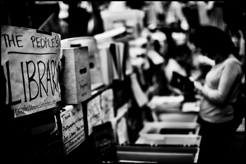 blurry photo of someone reading at an outdoor library; handwritten The People's Library sign in forefront