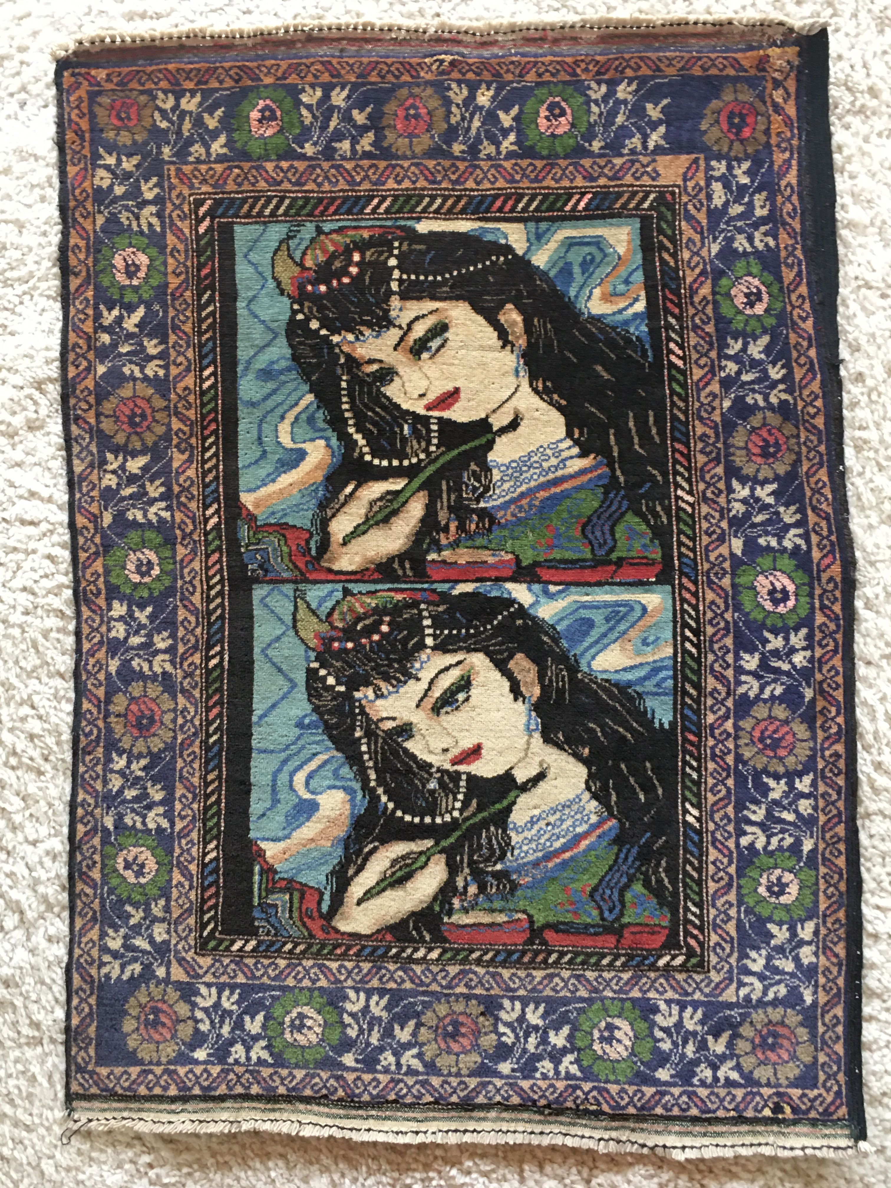 A full view of the Shirin rug. A complicated border of flowers and stripes frames two identical representations of the Princess Shirin. A hand is painting her as a portrait, with detailed facial features, long black hair, an embroidered green dress, and jewels. Behind the face in the portrait are zig-zagging lines that represent milk flowing down a mountain