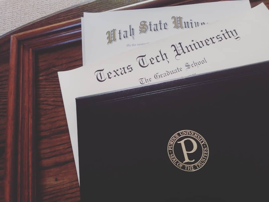 photograph of three diplomas layered on top of an empty wooden frame, one each from Utah State University, Texas Tech University, and Purdue University.