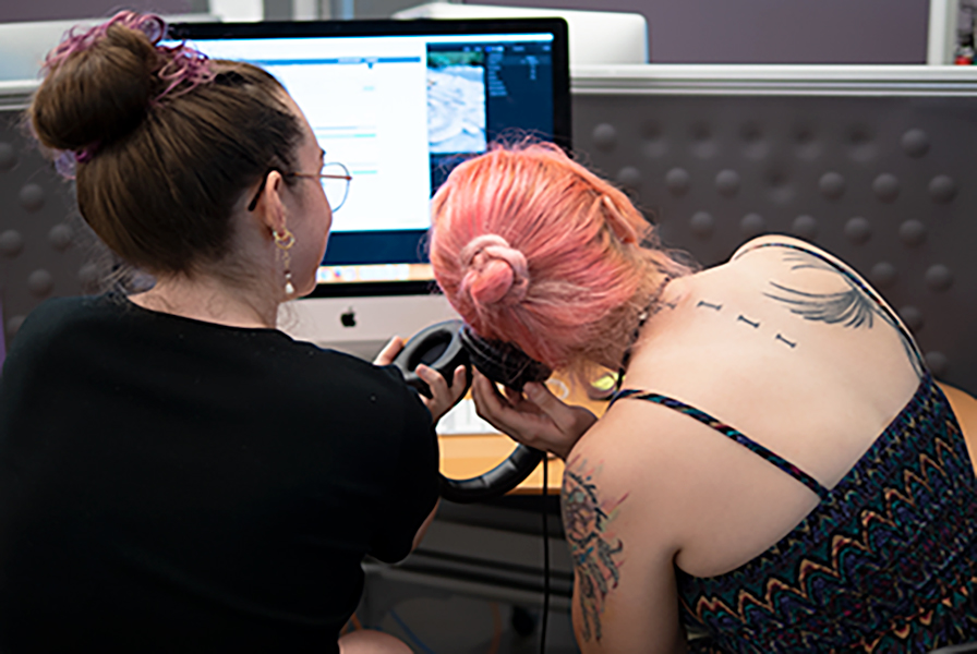 2 students working closely together at a computer, one leaning over to listen to headphones not on her head