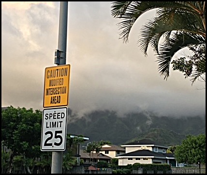 Light pole with posted speed limit sign of 25 MPH and yellow CAUTION sign that reads, modified intersection head. In the background are mist-covered mountains overlooking houses and palm trees.