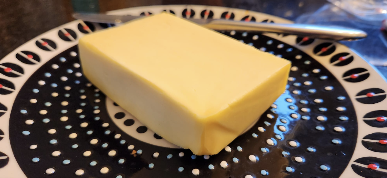 A block of butter positioned in the middle of a plate. A knife rests on the plate behind the block of butter.
