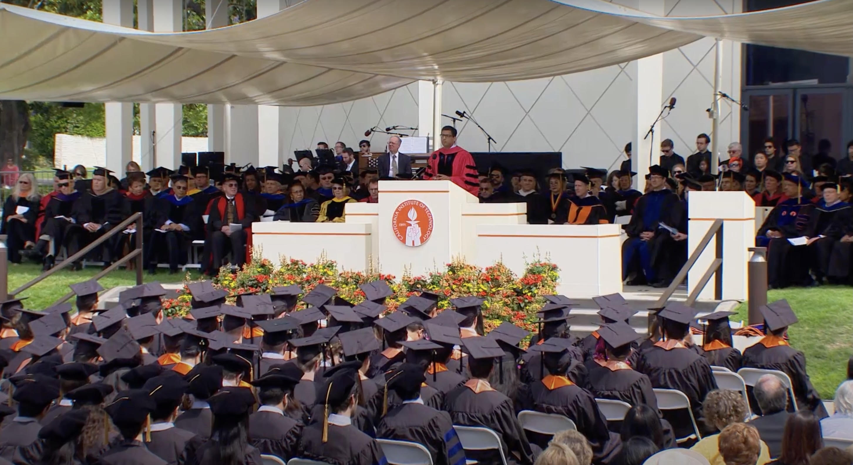 Atul Gawande delivering a commencement speech at a grand graduation podium. Multiple rows of graduates in black regalia are seated in the foreground, and two rows of faculty are seated onstage behind Gawande.