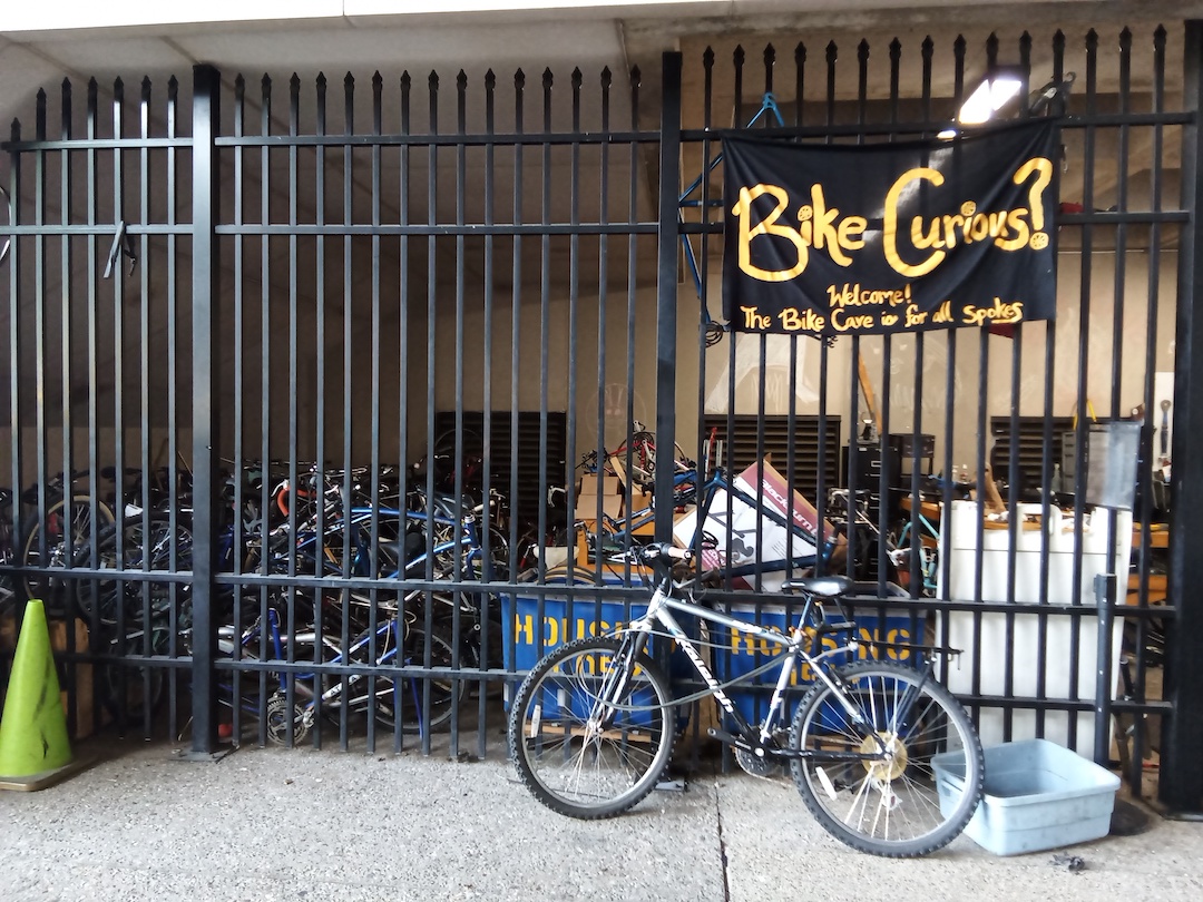 A bicycle leans against a large black fence with a sign that says, Bike Curious?