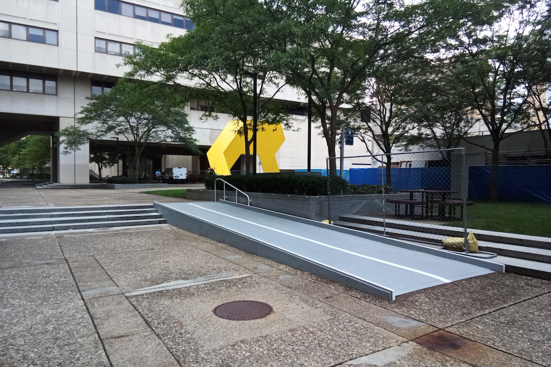 An empty courtyard with a set of stairs next to a metal ramp leading to a large concrete building.