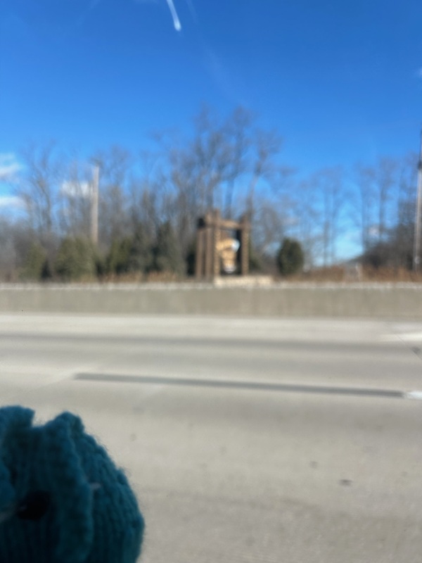 A blurry blue knitted bunny in front of a blurry Welcome to Wisconsin road sign.