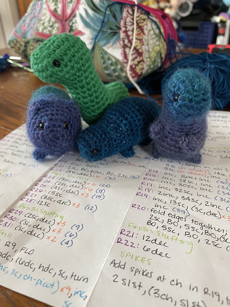 A group of blue, green, and purple crochet dinosaurs pose above printed out crochet patterns.