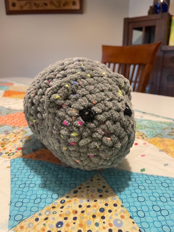 A gray crochet cat sits on a white, blue, and yellow quilted table runner.
