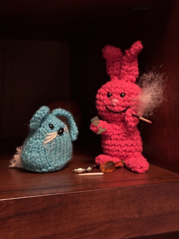 Evil Bunny holds a green wine glass in his right hand and is bringing a cigarette up to his mouth with his left. CC, the blue bunny, stands to Evil Bunny's left, watching him.