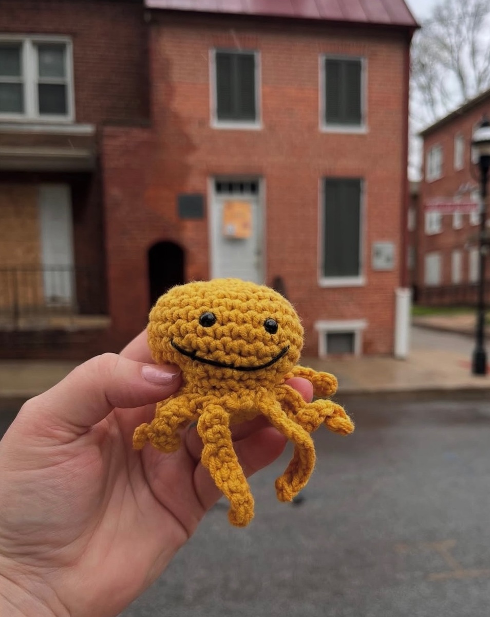 A smiling yellow crochet octopus is held in front of a row of red brick houses.