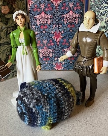 A blue, black, and green cat-like crochet animal sits on a beige table in the middle of plastic figurines of Jane Austen and William Shakespeare.