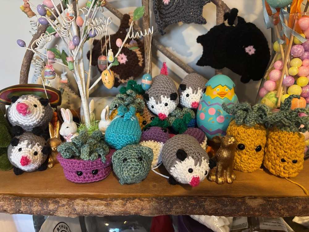 Crochet opossums, plants, pineapples, radishes, and cats are grouped on top of a wooden table among various Easter decorations.