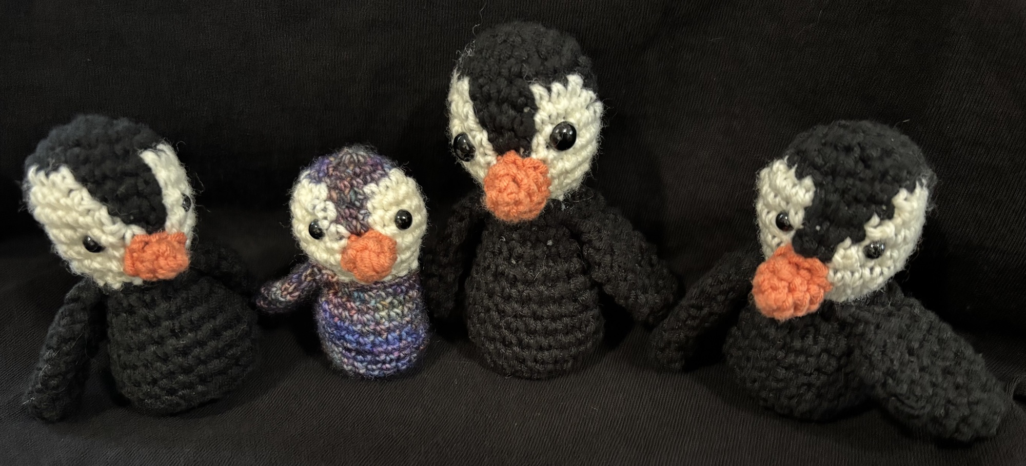 Four crochet penguins forming a half-circle and facing the camera. The penguin farthest to the left is the smallest and made with blue and purple speckled yarn. The other three penguins are black and white.