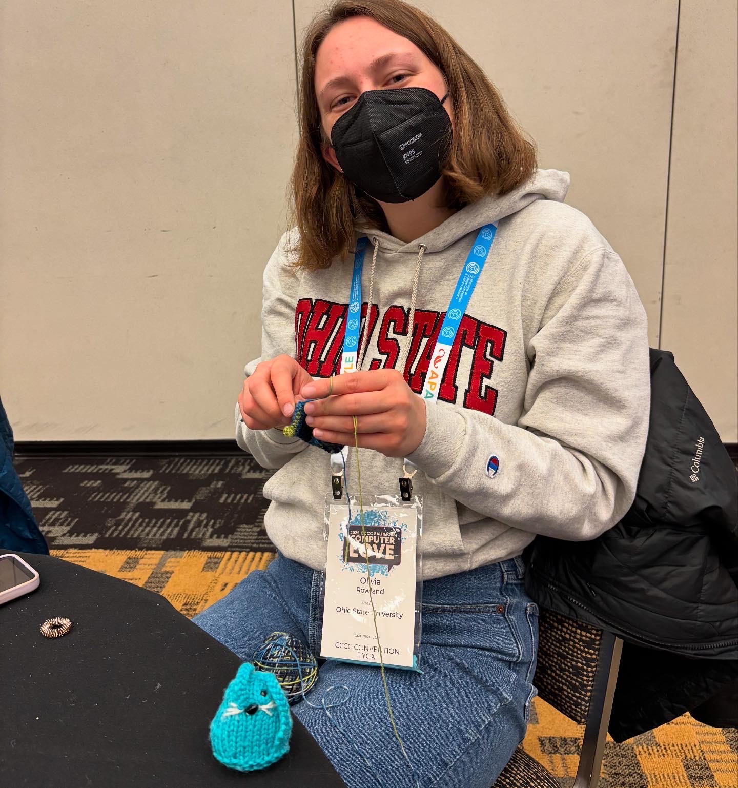 Olivia sits and crochets a conference creature. A small blue knit bunny rests on the table with a black tablecloth in front of her.