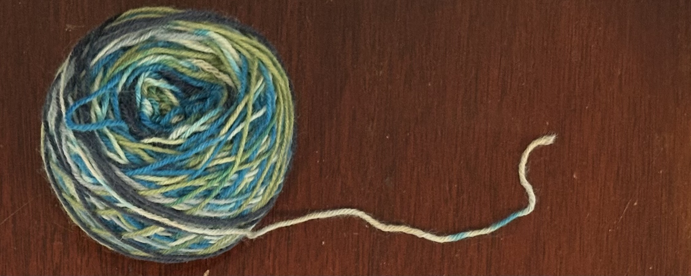 A ball of green, blue, and white yarn with the loose tail trailing to the right.