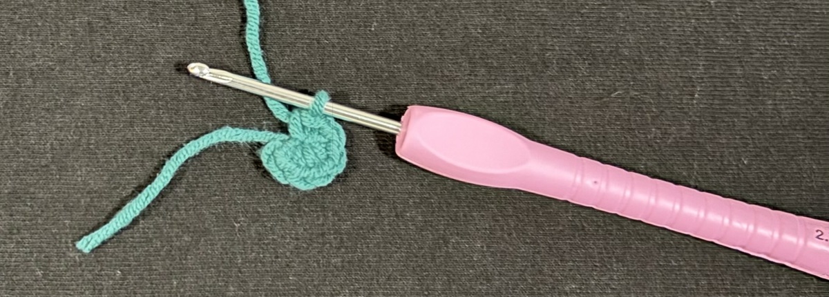 Banner A purple crochet hook with a small circle of crocheted blue yarn on top of a black background.