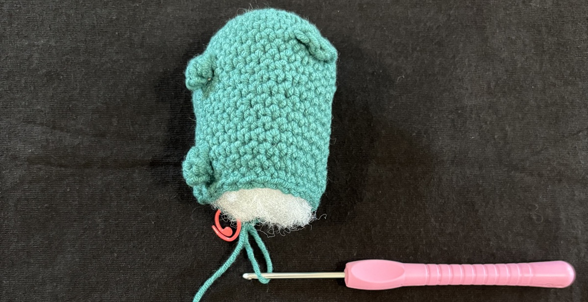 A blue crochet cat with white stuffing sticking out of the unfinished back attached to a purple crochet hook on a black background.