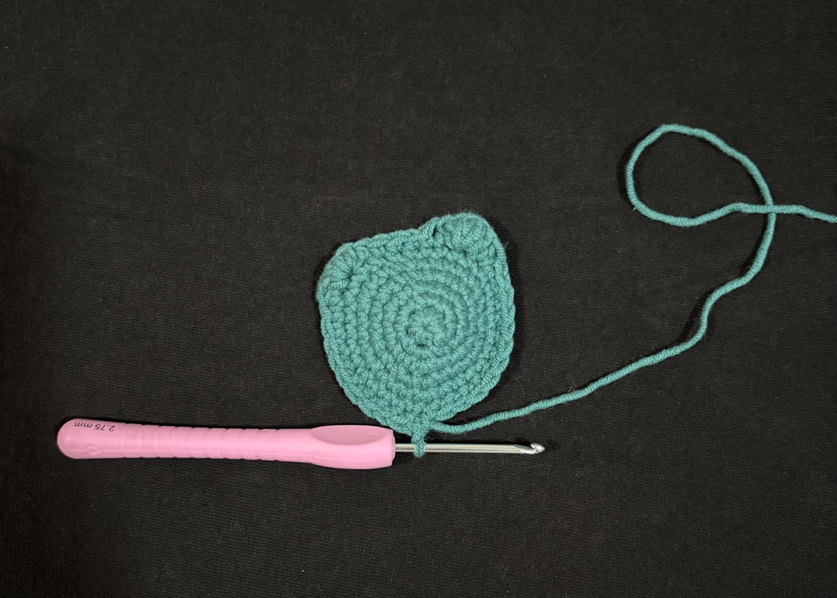 Blue crochet in the shape of a cat's face attached to a purple crochet hook on a black background.