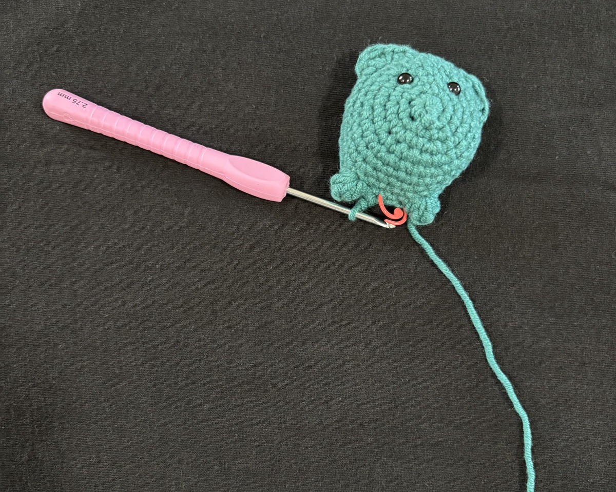 The front half of a blue crochet cat attached to a purple crochet hook on a black background.