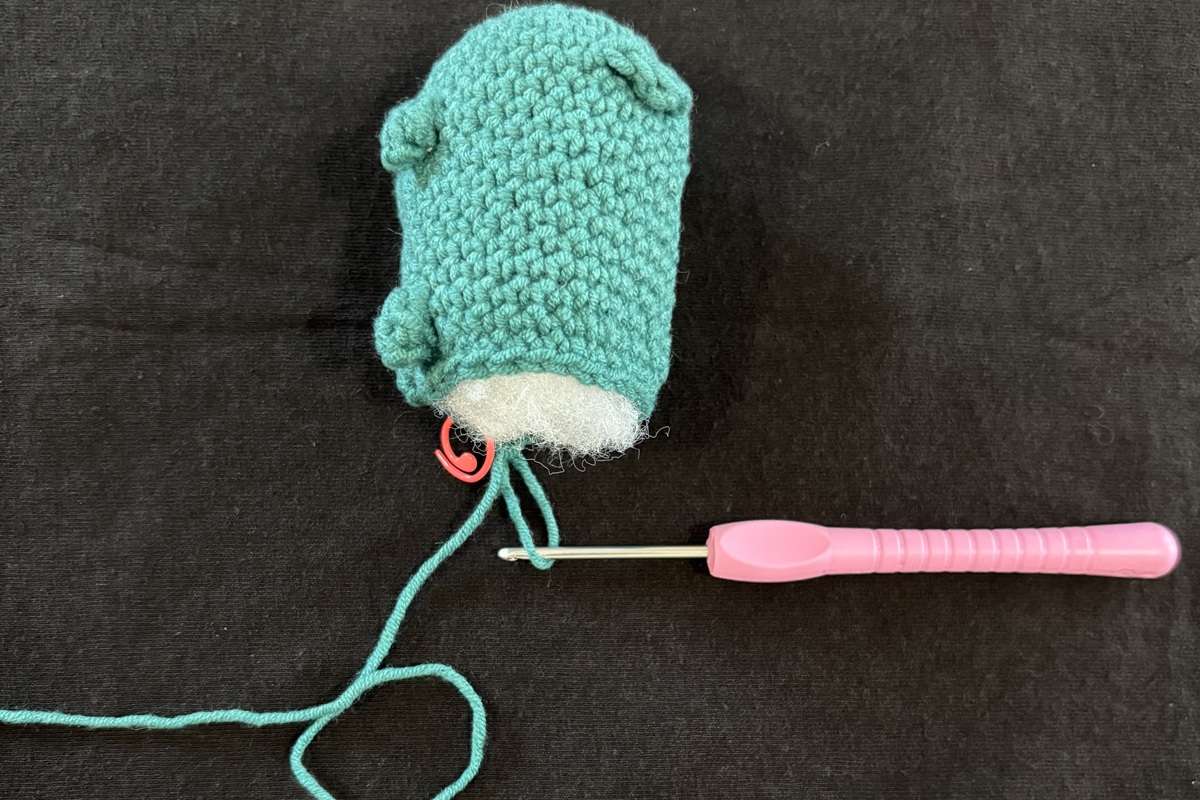 A blue crochet cat with white stuffing sticking out of the unfinished back attached to a purple crochet hook on a black background.