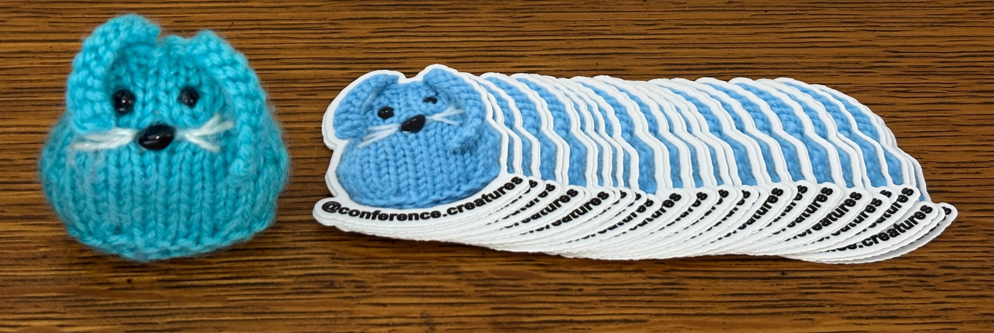 The blue knitted bunny Conference Creatures mascot, CC, standing to the left of a fanned-out pile of CC stickers that say 
